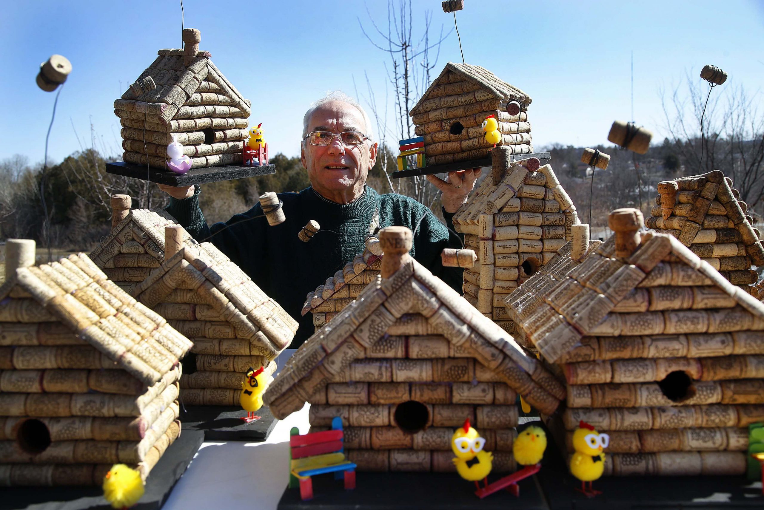 A man stands behind a table of birdhouses holding two birdhouses in the air